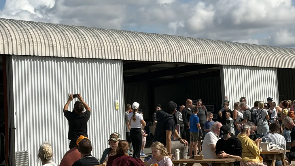 A visitor holding up a phone to photograph the fly-past, with crowds outside the hangar