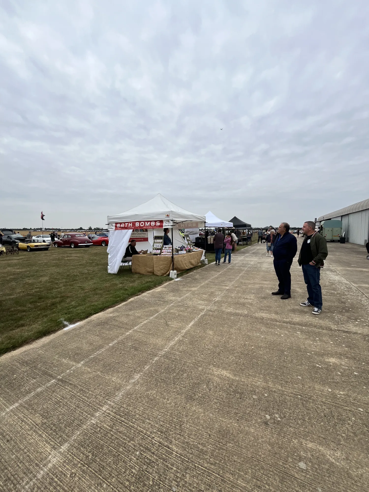 The concrete hardstanding at the hangar frontage running alongside the grass showground, with visitors walking between the stalls